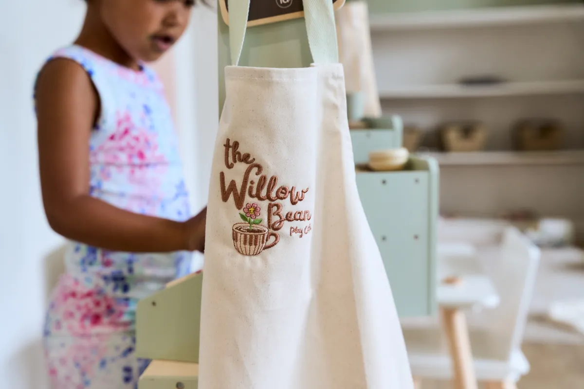 A child-sized apron embroidered with The Willow Bean Play Cafe logo, hanging at the children's play kitchen